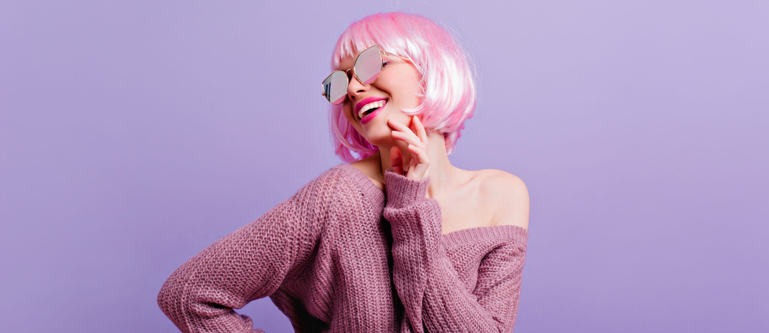 Pleased young girl in periwig and sunglasses having fun in studio. Photo of wonderful female model with pink hair smiling while dancing on purple background.